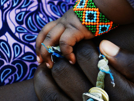 Close-up of a baby's hand holding an adult's finger, with a colorful beaded bracelet over purple patterned fabric.