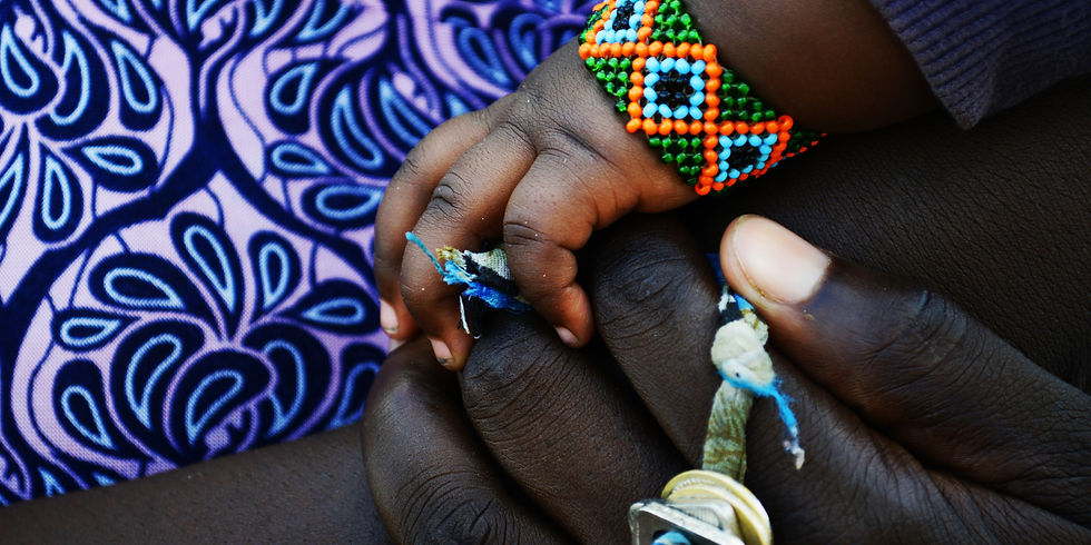 Close-up of a baby's hand holding an adult's finger, with a colorful beaded bracelet over purple patterned fabric.