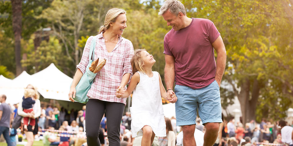 Smiling parents and daughter walk hand in hand at an outdoor park festival, with white tents and a crowd in the background.