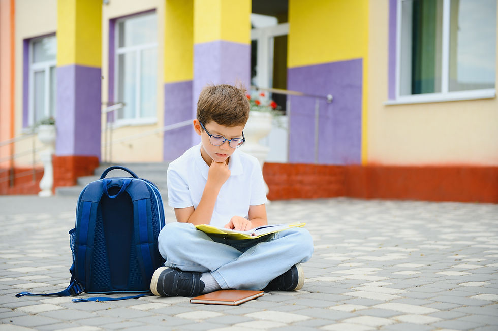 Boy with glasses sits cross-legged reading a book beside a blue backpack outside a colorful yellow and purple building.