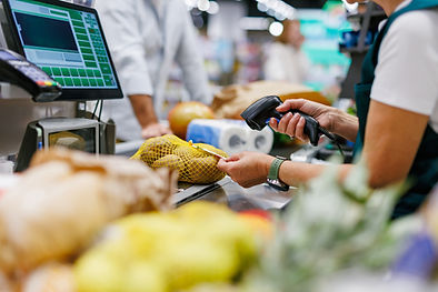 Grocery cashier scans a yellow mesh bag of potatoes at a checkout counter, with a green screen interface and shoppers blurred behind.
