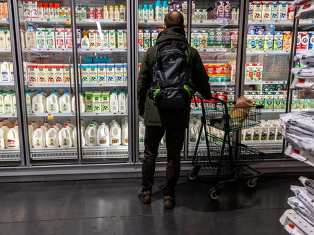 Man with backpack stands by a grocery cart in a dairy aisle, facing glass doors filled with milk cartons and cream.