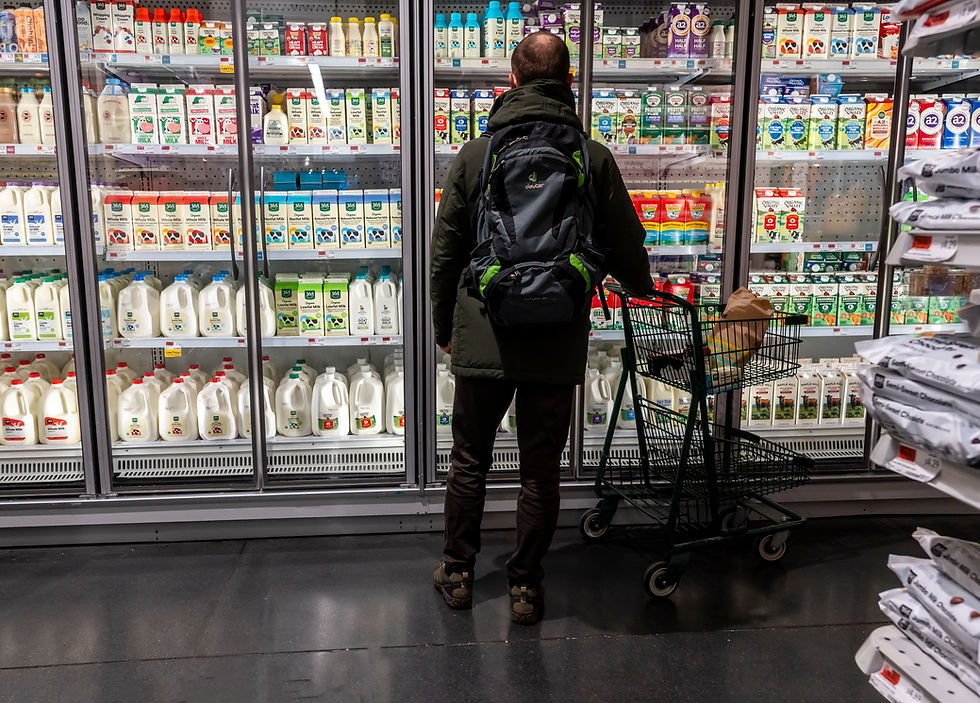 Man with backpack stands by a grocery cart in a dairy aisle, facing glass doors filled with milk cartons and cream.