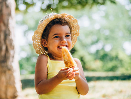 Smiling child in a straw hat holds a popsicle outdoors, wearing a yellow top against a soft green background.