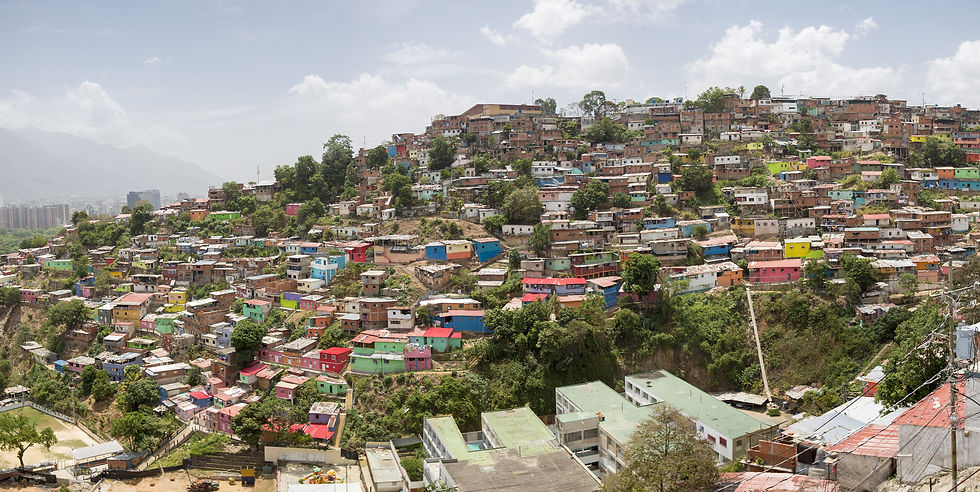 Colorful hillside favela with densely packed homes, green trees, and distant city under a bright cloudy sky.
